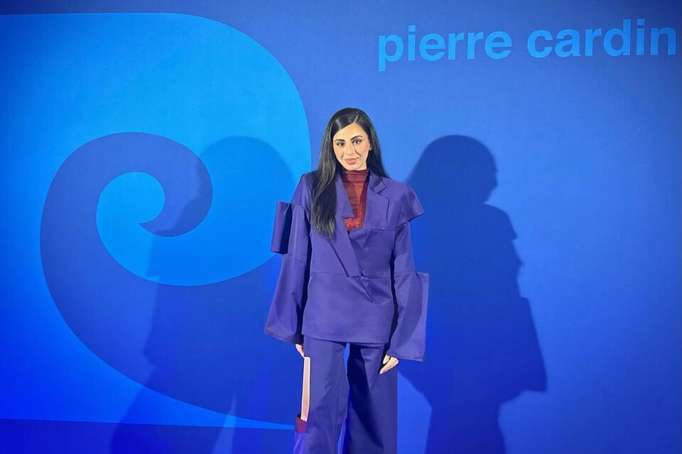 פרלה סיבני עומדת ומאחוריה רולאפ עם המיתוג של PIERRE CARDIN